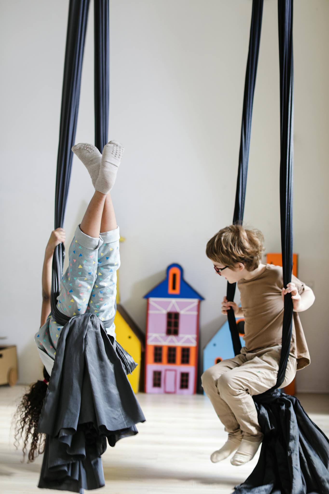 Two children having fun swinging indoors with colorful playhouses in the background.